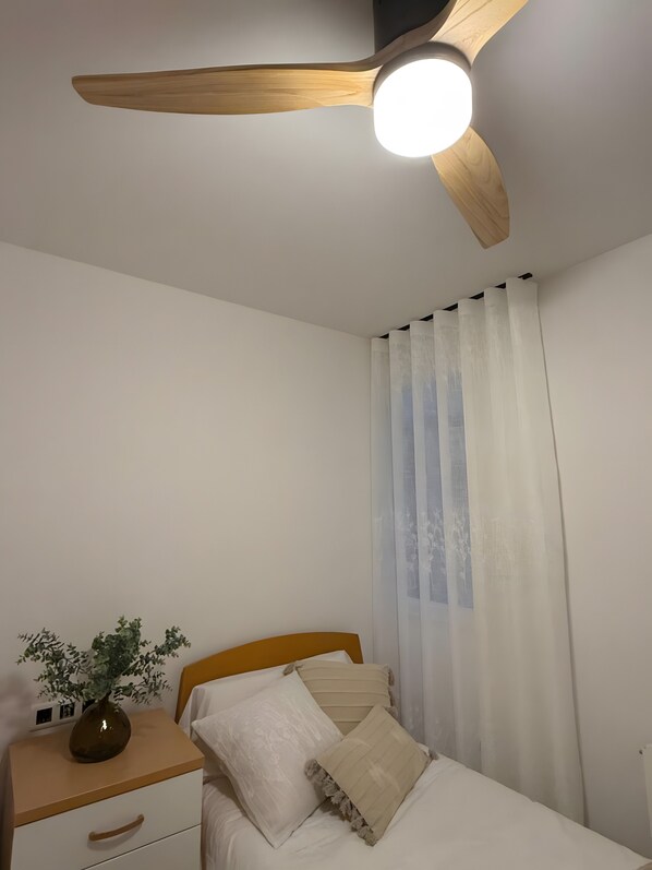 Ein Schlafzimmer mit einem Bett, einem Nachttisch, einer Pflanze in einer Vase, einem Deckenventilator und leichten Vorhängen.