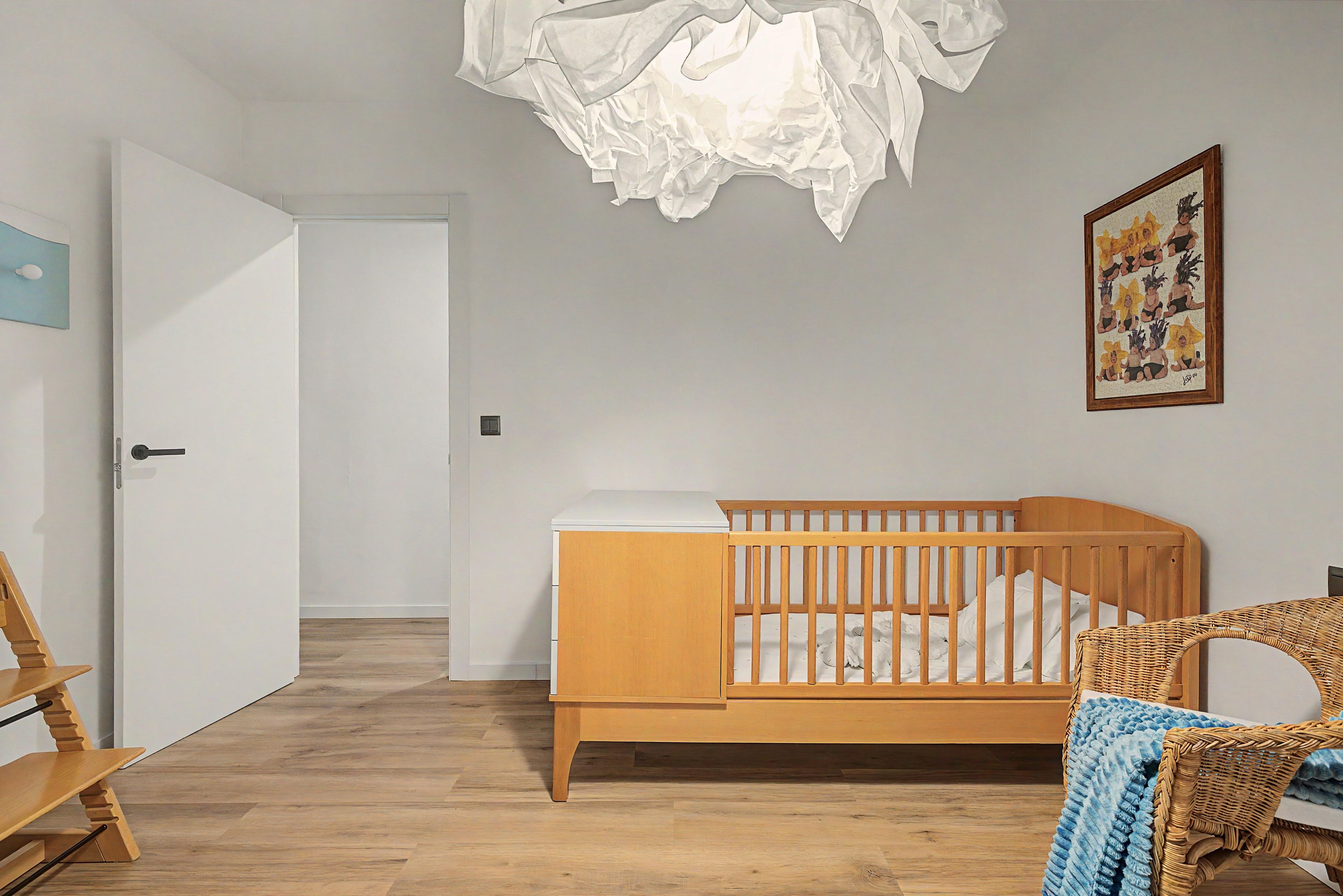 Una cuna para bebé con estructura de madera y colchón blanco, una silla de mimbre y un cuadro enmarcado en la pared.