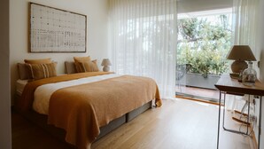Ein Schlafzimmer mit einem großen Bett, einem an der Wand befestigten Kunstwerk, einem Tisch mit einer Lampe und Blick auf Grünflächen.