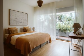 Een slaapkamer met een groot bed, een aan de muur hangend kunstwerk, een tafel met een lamp en uitzicht op groen buiten.