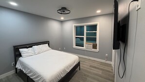 Un dormitorio con cama, ventana, ventilador de techo y televisor montado en la pared.