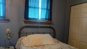 A bedroom with a bed, a bedside table, a window with blinds, and a door.