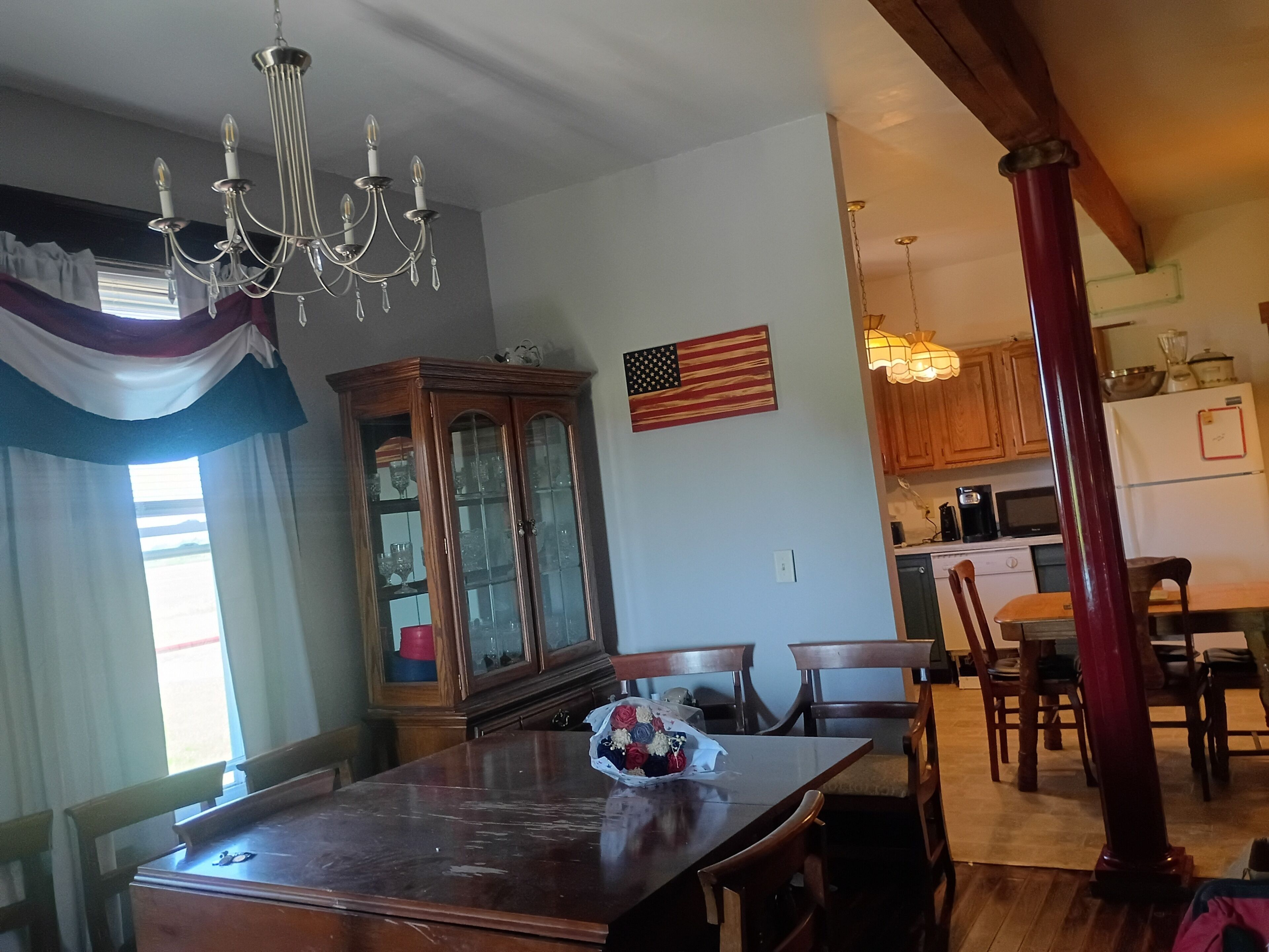 Zona pranzo con tavolo in legno, sedie, un lampadario, un armadio con vetrate e una cucina sullo sfondo.