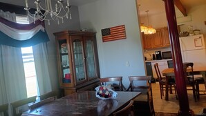 A dining area with a wooden table, chairs, a chandelier, a cabinet with glass doors, and a kitchen in the background.