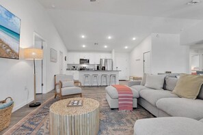 Ein modernes Wohnzimmer mit Sofa, Sesseln und Couchtisch, im Hintergrund eine Küche und ein großes Bild an der Wand.