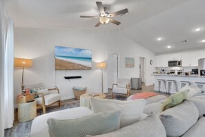 Ein modernes Wohnzimmer mit einer großen Couch, einem Couchtisch und einem an der Wand befestigten Fernseher. Es gibt einen Deckenventilator, eine Küchenzeile mit weißen Schränken und einen Essbereich mit Barhocker.