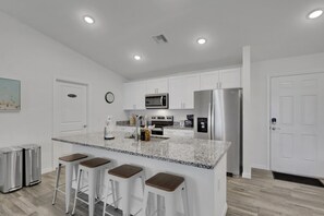 A modern kitchen with white cabinets, stainless steel appliances, a granite countertop, and bar stools. - Modern Cape Coral Oasis with Saltwater Pool! (Cape Coral)