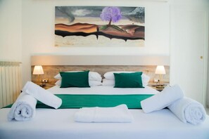 Habitación de hotel con una cama, almohadas verdes, ropa de cama blanca, dos lámparas de noche y un cuadro sobre la cama.