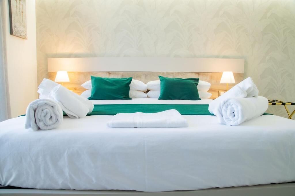 Una cama bien hecha con ropa de cama blanca y verde, dos lámparas de noche y un cabecero con una lámpara.