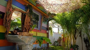 Un bâtiment aux couleurs vives, agrémenté d’une fontaine en pierre, de plantes tropicales et d’une structure en bois.