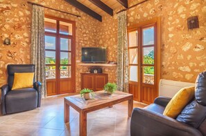 A cozy living room with a stone-patterned wall, a wooden coffee table, a TV, and a view of greenery outside. - Son Julia de sa Torre by Interhome (Porreres)