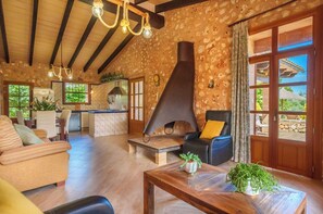 A spacious living area with a stone wall, wooden ceiling beams, a large fireplace, and a view of the outdoors through a glass door. - Son Julia de sa Torre by Interhome (Porreres)