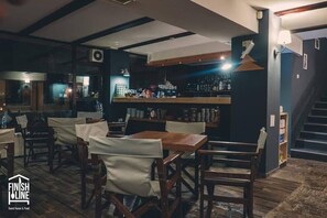 Un restaurante con mesas y sillas de madera, una zona de bar con estanterías llenas de botellas y una escalera.