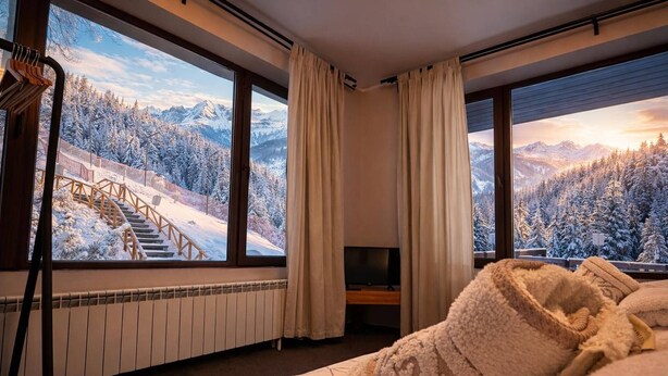 A hotel room with a large window offering a view of a snowy mountain landscape, a bed with a patterned bedspread, and a television on a stand. - Finish Line Guest House (Samokov)