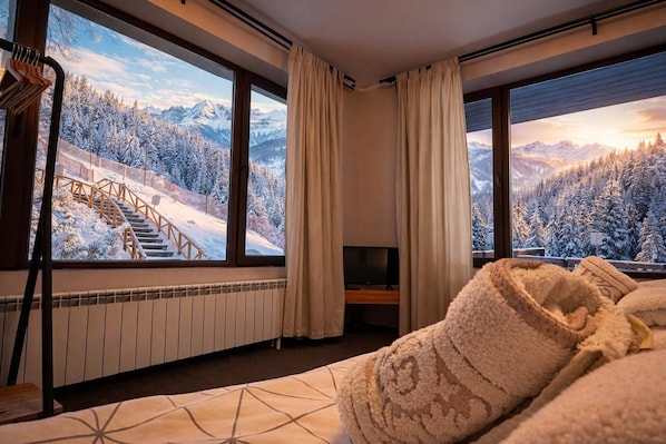Habitación de hotel con un ventanal grande que ofrece vistas a un paisaje montañoso nevado, una cama con colcha estampada y un televisor sobre un soporte.