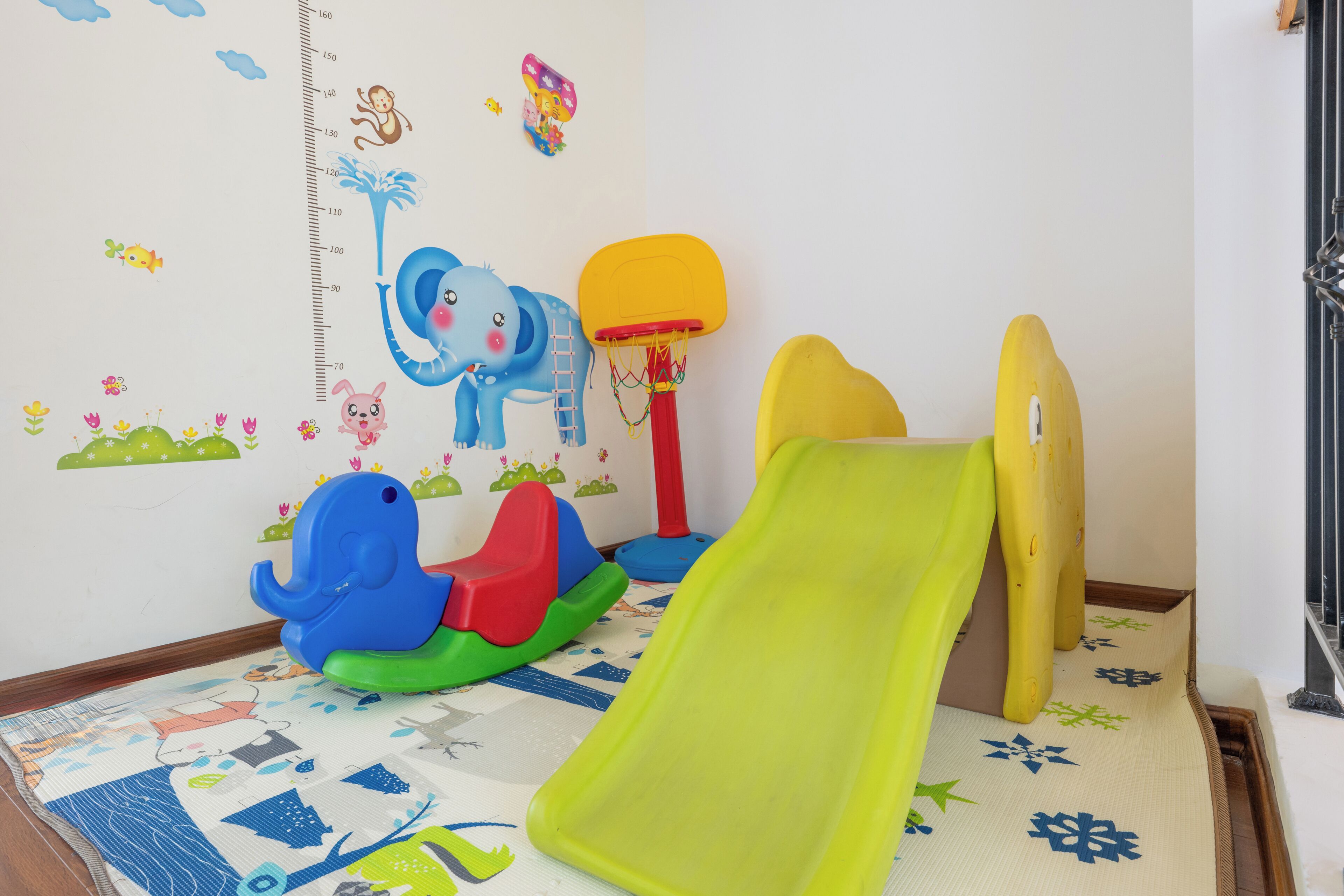 Zona infantil con un tobogán, un elefante de juguete para trepar y una pared con una tabla de medidas de altura y pegatinas de dibujos animados.
