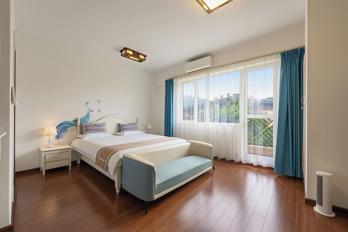 Ein Schlafzimmer mit Bett, Nachttisch, Lampe, Klimaanlage, Fenster mit Vorhängen und Blick nach draußen.