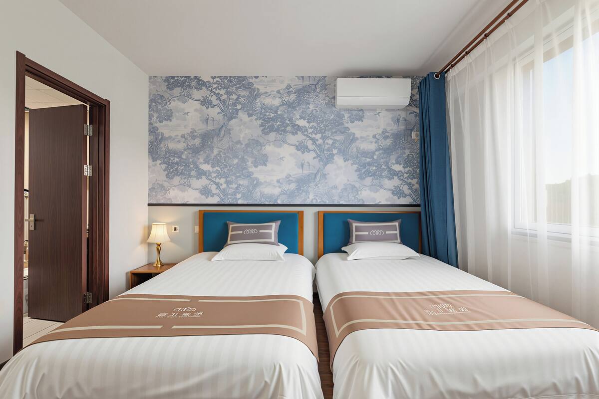 Ein Hotelzimmer mit zwei Betten, einem Nachttisch mit Lampe, einer Wand-Klimaanlage und einem Fenster mit Vorhängen.
