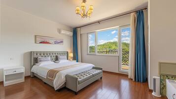 A bedroom with a bed, bedside tables, a bench, a chandelier, a window with curtains, and a view of greenery.