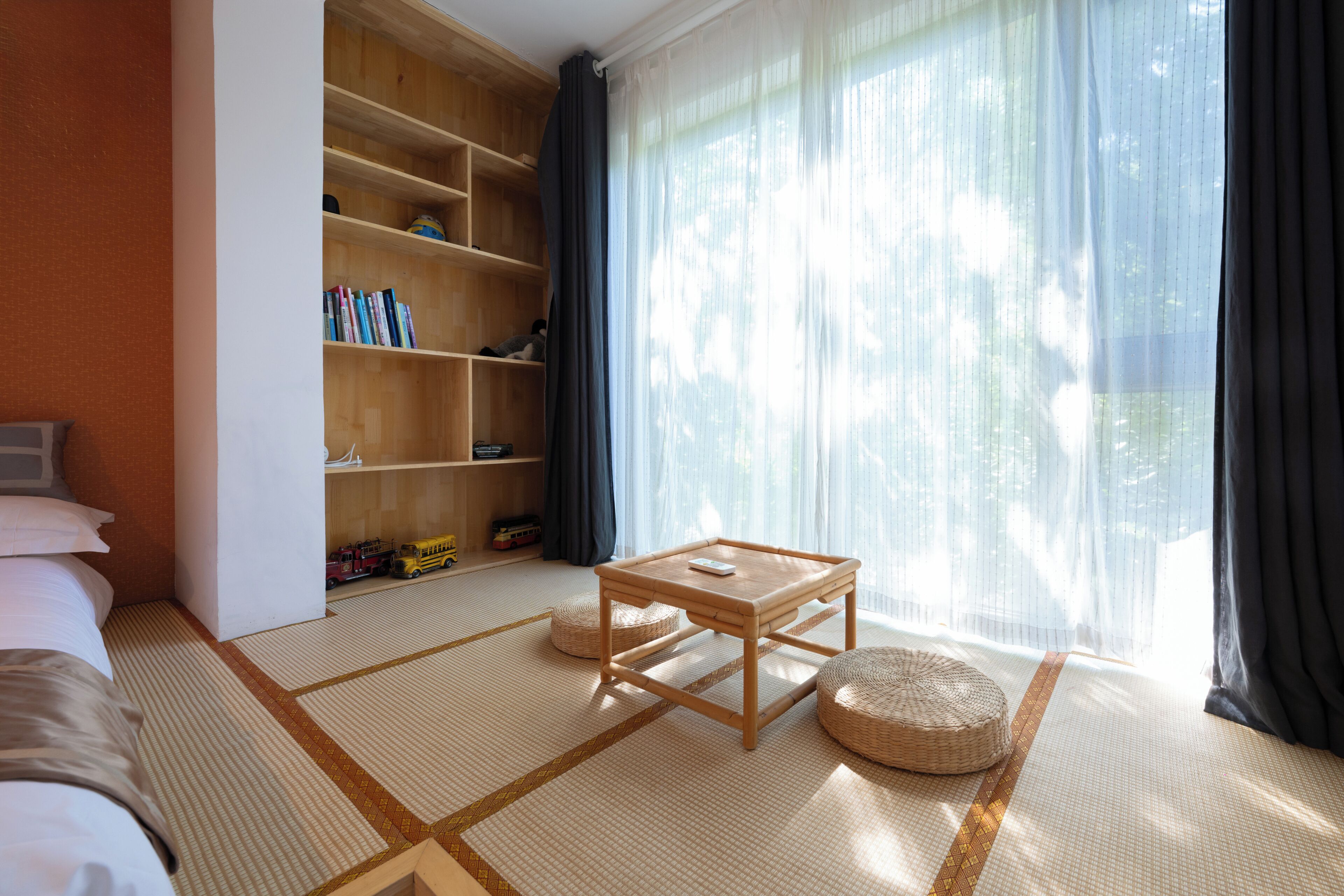 Una habitación con una cama, una estantería de madera, una mesita y vistas al exterior a través de cortinas traslúcidas.
