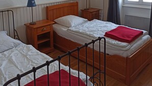 Een kamer met twee houten bedden, een nachtkastje met een lamp, een wandplank met decoratieve objecten, en een raam met gordijnen.