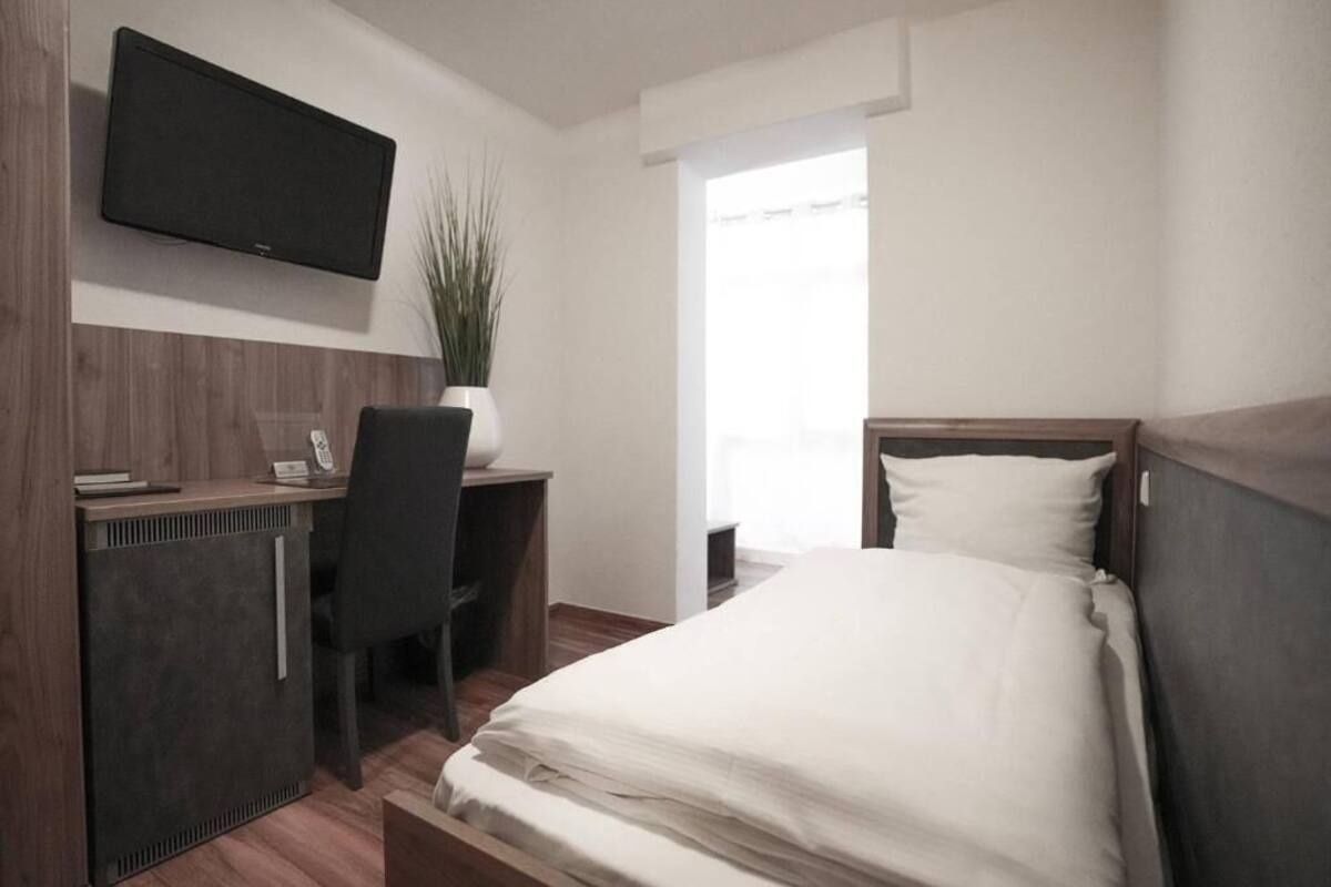 A hotel room with a bed, a desk with a chair, a television mounted on the wall, and a window with curtains.
