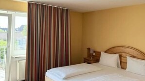A double bed with a wooden headboard, a bedside table, and a window with curtains. - Hotel Altavilla (Schwetzingen)