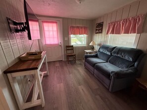 A cozy room with a wooden floor, a blue sofa, a small table, and a door with pink curtains. - Mangrove Landing - Debbie’s Roseate (Cedar Key)