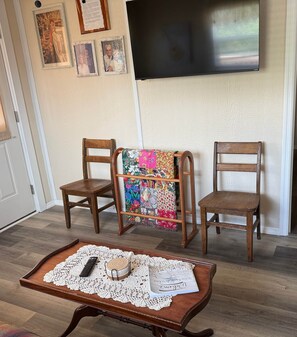 Ein Wohnzimmer mit zwei Holzstühlen, einem kleinen Tisch mit Spitzenunterlage, einer Steppdecke und einem flachbildfernseher an der Wand.