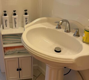 A bathroom with a white sink, two faucets, and three bottles of toiletries on a shelf. - Mangrove Landing - High Tide (Cedar Key)