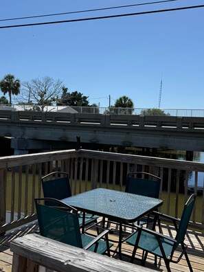 Eine Holzterrasse mit Tisch und Stühlen, mit Blick auf ein Gewässer und eine Brücke.