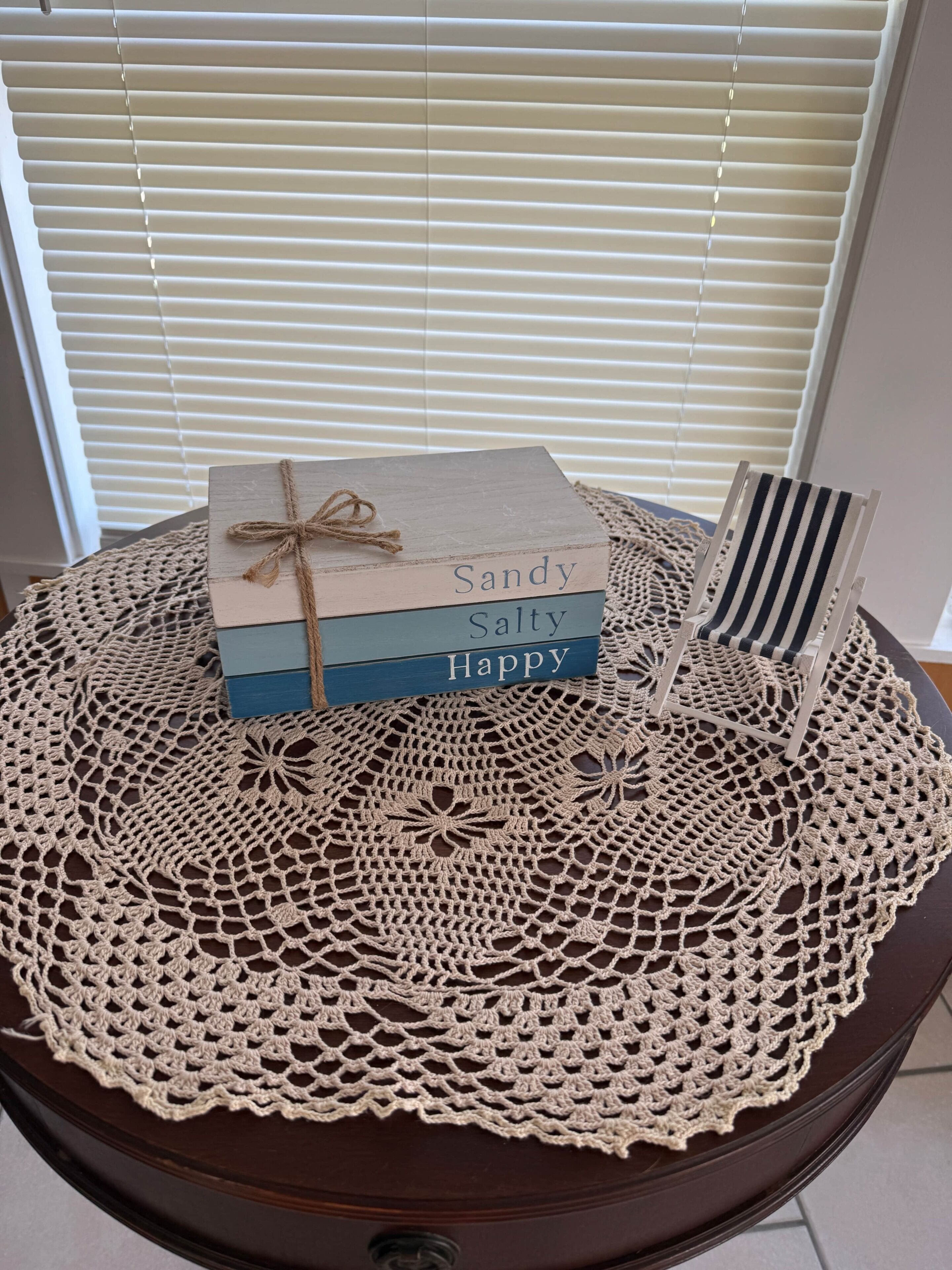 A table with a crocheted tablecloth, a small wooden box with the name 'Sandy Salty Happy' tied with a string, and a striped chair.