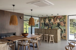 Ein moderner Essbereich mit einem Holztisch, Stühlen und einer großen Schüssel. Es gibt Pendelleuchten und ein Wandbild mit tropischen Blättern.