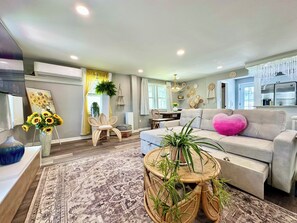 Ein Wohnzimmer mit einer Couch, einem runden Couchtisch, einem Teppich, einem Sessel und einer Essecke im Hintergrund.