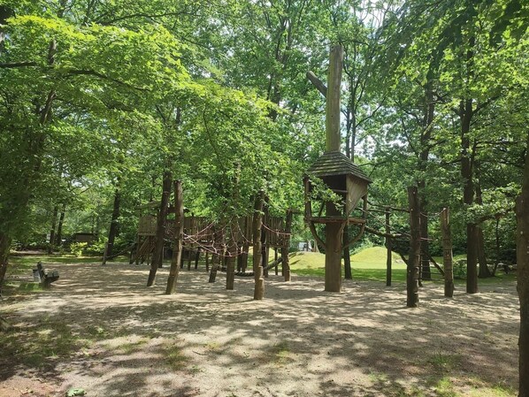 Un parque infantil con estructuras para escalar y una casita de madera en un área boscosa.