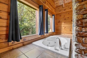 Interno di una baita in legno con una grande vasca, pareti in legno e vista sugli alberi dalle finestre.