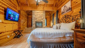 A wooden cabin interior with a stone fireplace, a bed with white bedding, a television mounted on the wall, and a ceiling fan.