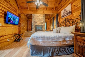 Ein Holzblockhaus-Interieur mit Kamin aus Stein, einem Bett mit weißer Bettwäsche, einem an der Wand befestigten Fernseher und einem Deckenventilator.