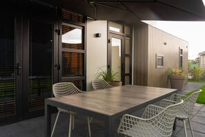 An outdoor dining area with a wooden table and white chairs, a potted plant, and a modern building with wooden siding. - Prachtig luxe afgewerkt en splinternieuw chalet (Molenschot)
