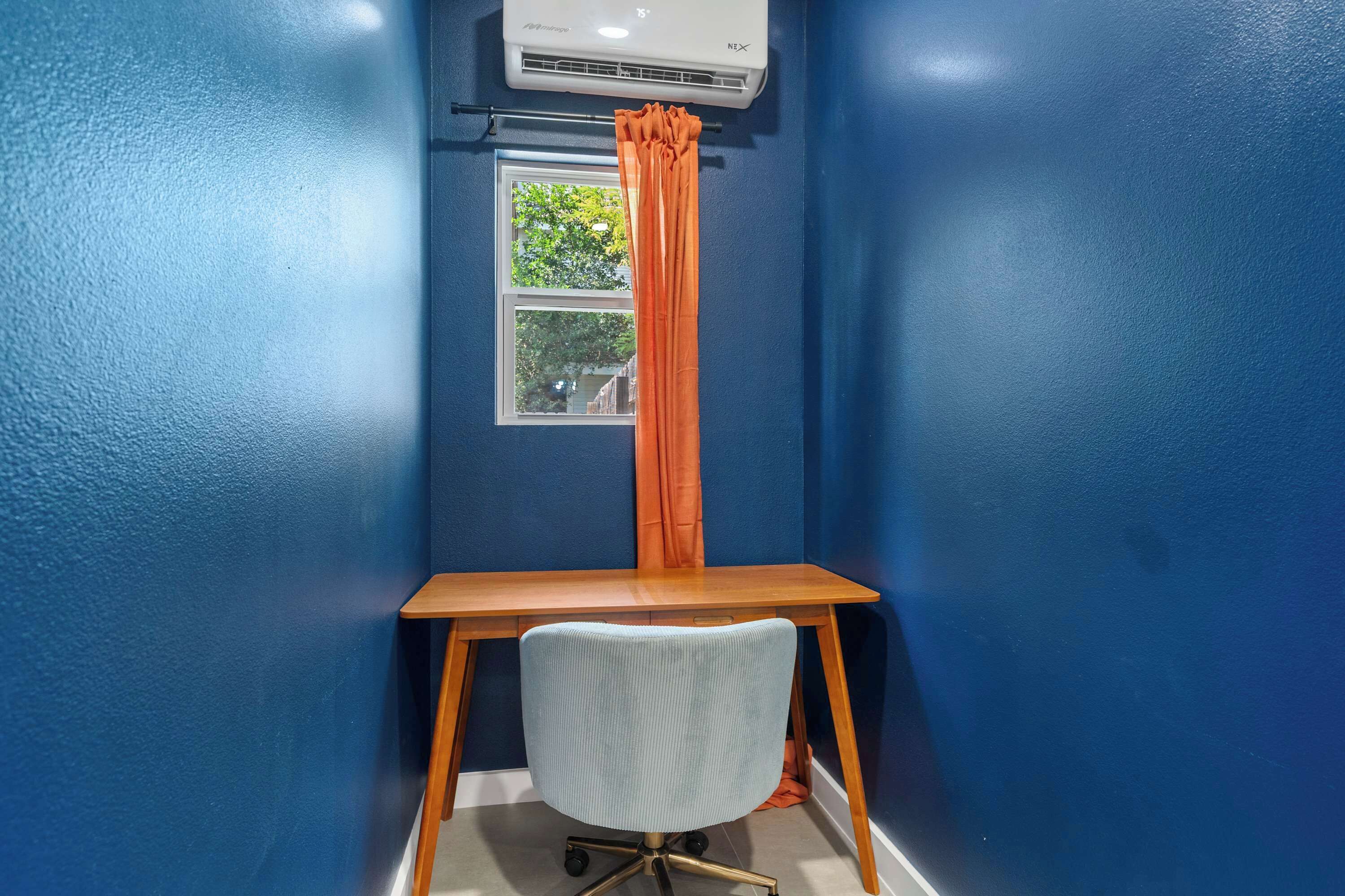 Een kamer met blauwe wanden, een houten bureau, een witte stoel en een airconditioning boven een raam met een oranje gordijn.