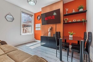Un salon moderne avec une table à manger en verre, des chaises noires, un téléviseur fixé sur un mur orange et une fenêtre donnant sur un immeuble.
