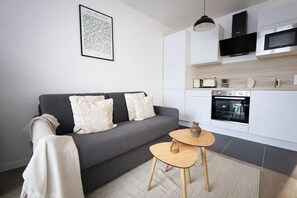 Ein modernes Wohnzimmer mit einem grauen Sofa, einem kleinen runden Holztisch und einer Küchenzeile mit weißen Schränken und Geräten.