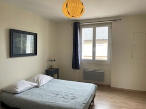 Une chambre à coucher avec un lit, une table de chevet, une lampe, une fenêtre avec des rideaux, un radiateur et un tableau encadré au mur.
