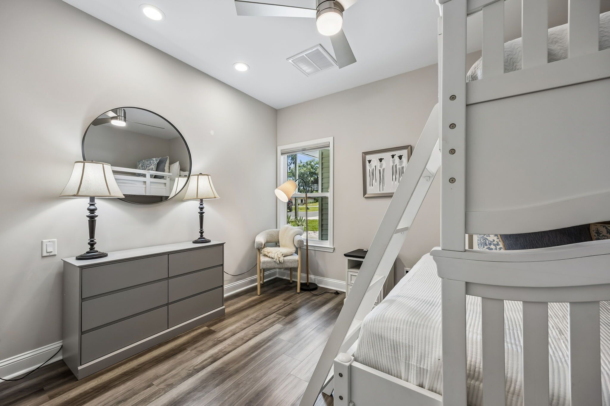 A bedroom with a bunk bed, a dresser, a chair, a round mirror, and a window.