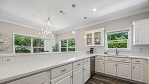 Une cuisine moderne avec des meubles blancs, un grand îlot central et de nombreuses fenêtres offrant une vue sur la verdure.