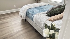 Un dormitorio con una cama, una mesita de noche, un florero con flores y dos ventanas.