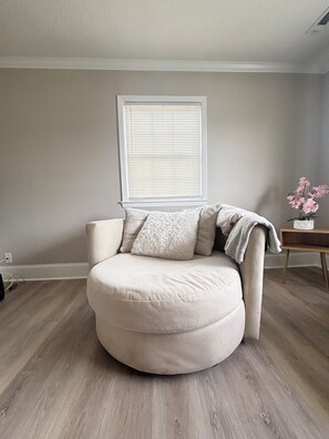 A round, beige armchair with a fluffy pillow and a blanket, positioned in a room with light wooden flooring, a white wall, and a window with blinds. - Mins from Duke Hospital and UNC Chapel Hill (Durham)