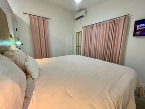 A bedroom with a bed, curtains, an air conditioning unit, a television, and a wall-mounted lamp.