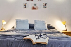 Una cama con un libro y gafas, dos lámparas de noche y una pared con obras de arte enmarcadas.
