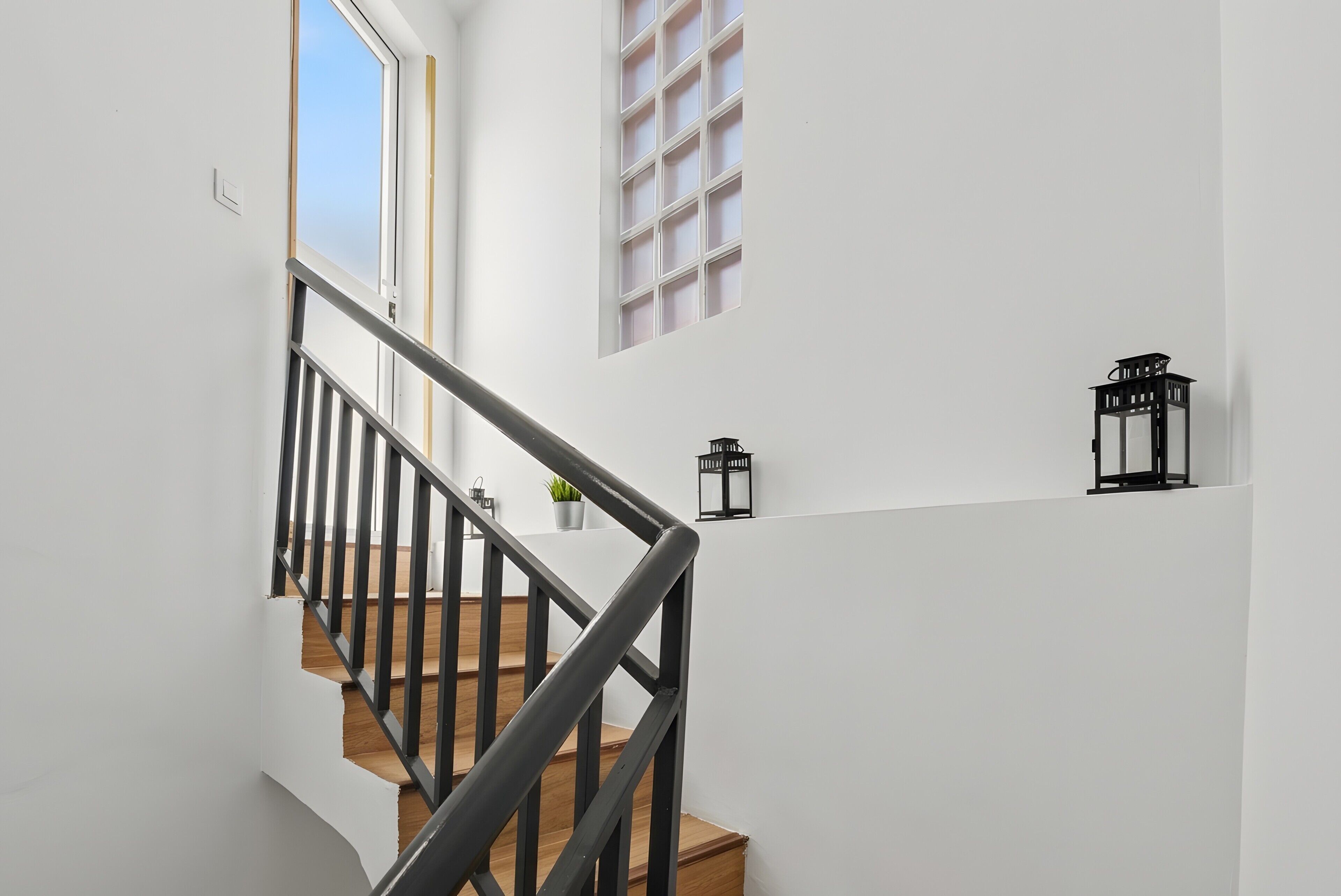 Una escalera con barandillas negras y detalles de madera, que conduce a una plataforma con una planta en maceta y un farol negro.